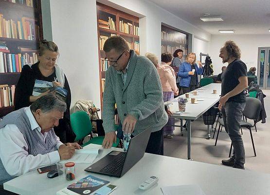 Mężczyzna siedzący przy stole podpisuje egzemplarz książki, przy stole stoi drugi mężczyzna i kobieta.