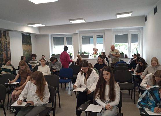Osoby siedzące na sali, przy krzesłach mają stoliki na których piszą test.