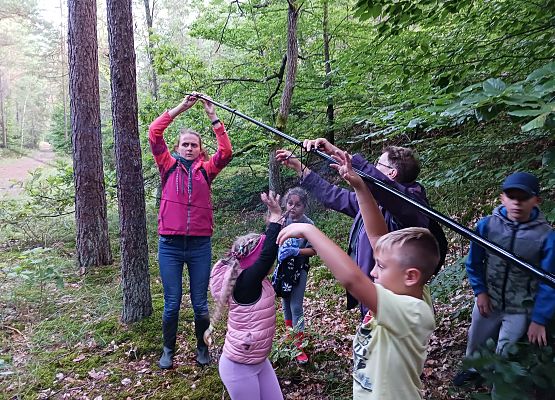 Dwie osoby dorosłe i kilkoro dzieci trzymają pod katem  wysokie tyczki na które jest zakładana cieniutka sieć.