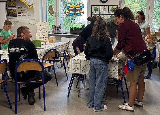 Dzieci siedzą przy stołach i malują, dorośli stoją za ich plecami.