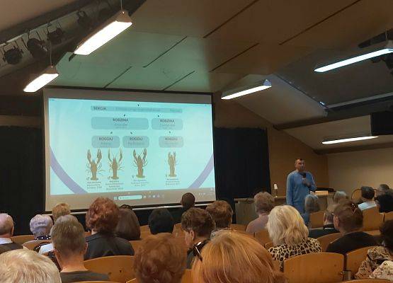 Na pierwszym planie głowy słuchaczy Uniwesytetu Trzeciego Wieku, w głębi prowadzący stoi przy ekranie  na którym są sylwetki 4 raków