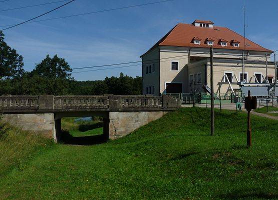 Duzy jasny budynek z czerwonym dachem, na pierwszym planie mały wiadukt pod drogą.
